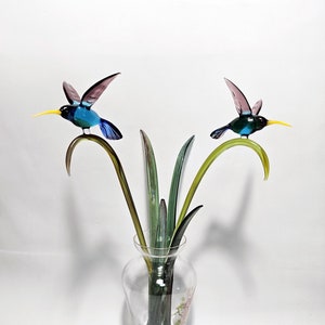 Könnte beinhalten: Eine Glasvase mit Blumenmuster enthält zwei Glas-Kolibri-Skulpturen mit grünen, blauen und violetten Flügeln. Die Kolibris sitzen auf grünen Glasstielen.