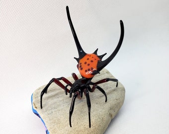 May include: A black and red glass spider sculpture with long legs and a spiky, orange and black patterned body. The spider is perched on a gray rock with a blue base.