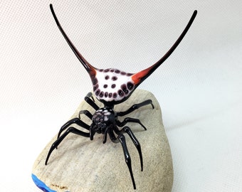 May include: A glass sculpture of a spider with a white and black body and red and black legs. The spider is perched on a small, gray rock with blue and purple accents.