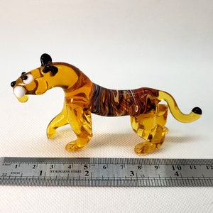 K&ouml;nnte beinhalten: Eine kleine Glas-Tigerfigur mit bernsteinfarbenen und braunen Streifen. Die Figur ist etwa 10 cm lang und hat schwarze Augen und eine schwarze Nase.