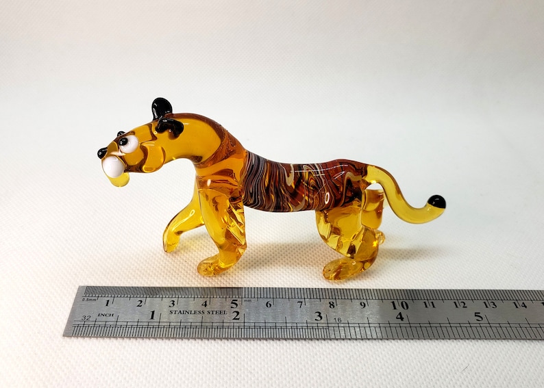 K&ouml;nnte beinhalten: Eine kleine Glas-Tigerfigur mit bernsteinfarbenen und braunen Streifen. Die Figur ist etwa 10 cm lang und hat schwarze Augen und eine schwarze Nase.