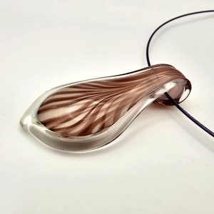 Puede incluir: Un colgante de vidrio transparente con un diseño de espiral marrón que cuelga de un cordón negro. El colgante tiene forma de lágrima y tiene un acabado liso y pulido.