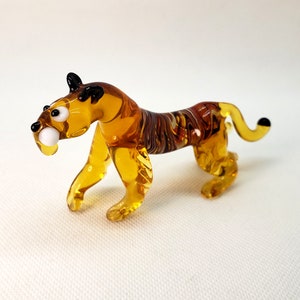K&ouml;nnte beinhalten: Eine kleine Glasfigur eines Tigers mit bernsteinfarbenen und braunen Wirbeln. Der Tiger hat schwarze Augen und einen wei&szlig;en Zahn.