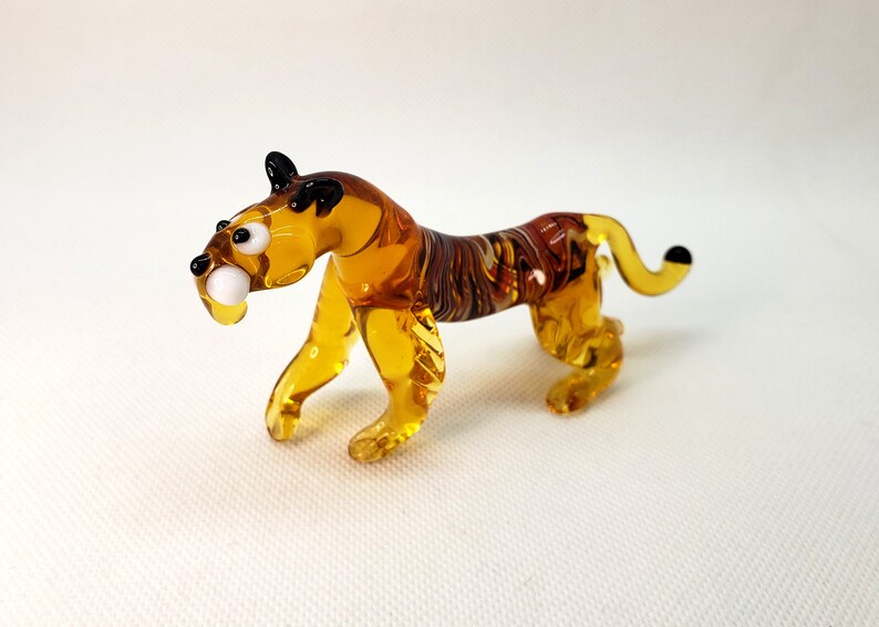 K&ouml;nnte beinhalten: Eine kleine Glasfigur eines Tigers mit bernsteinfarbenen und braunen Wirbeln. Der Tiger hat schwarze Augen und einen wei&szlig;en Zahn.