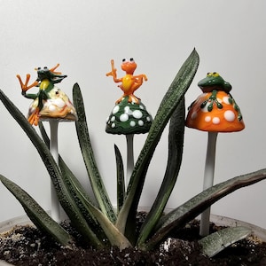 Puede incluir: Tres figuras de ranas de vidrio sobre tallos con forma de hongo, colocadas en una suculenta en maceta. Las ranas son de color verde, naranja y amarillo, con lunares blancos en los sombreros de los hongos. Un artículo de decoración único.