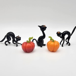 May include: Three black glass cats with orange and red glass pumpkins. The cats are in various poses, one is looking at a pumpkin.
