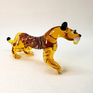 K&ouml;nnte beinhalten: Eine Glas-Tigerfigur mit bernsteinfarbenen und braunen Streifen. Die Figur hat schwarze Augen und einen wei&szlig;en Zahn.