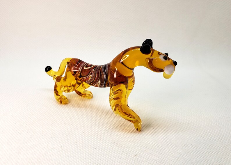 K&ouml;nnte beinhalten: Eine Glas-Tigerfigur mit bernsteinfarbenen und braunen Streifen. Die Figur hat schwarze Augen und einen wei&szlig;en Zahn.