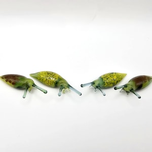 Może przedstawiać: Cztery szklane ślimaki w różnych odcieniach zieleni i żółci z brązowymi i niebieskimi akcentami. Ślimaki są ułożone w rzędzie na białym tle.