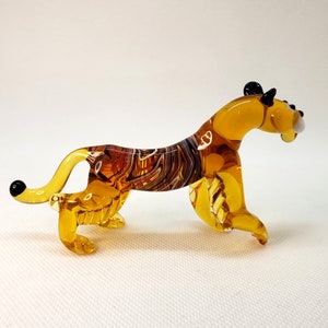 K&ouml;nnte beinhalten: Eine Glasfigur eines Tigers mit einem braun-bernsteinfarbenen gestreiften K&ouml;rper und schwarzen Ohren und Nase.