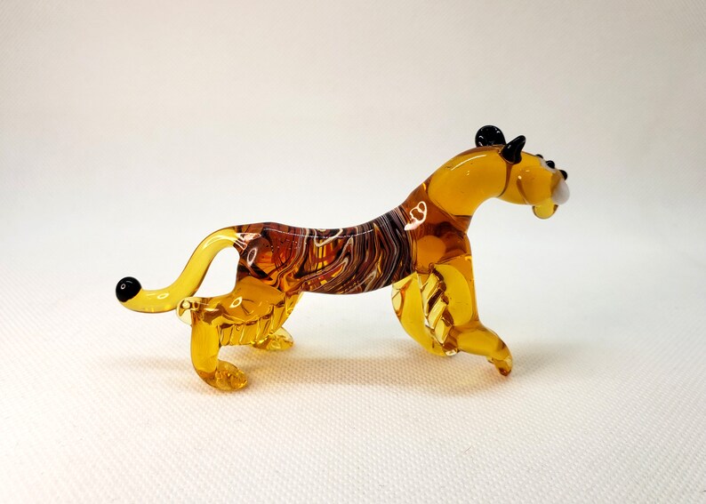 K&ouml;nnte beinhalten: Eine Glasfigur eines Tigers mit einem braun-bernsteinfarbenen gestreiften K&ouml;rper und schwarzen Ohren und Nase.