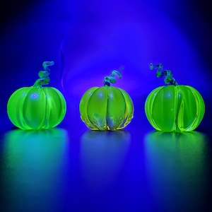 May include: Three glowing green glass pumpkins with curly stems are arranged on a reflective surface against a dark blue background. The pumpkins are illuminated, creating a vibrant and eye-catching display. Perfect for Halloween or fall decor.
