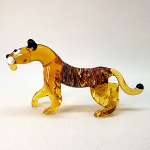 K&ouml;nnte beinhalten: Eine Glasfigur eines Tigers mit einem gelb-braun gestreiften K&ouml;rper, schwarzen Ohren und wei&szlig;en Augen. Die Figur steht auf einer wei&szlig;en Oberfl&auml;che.