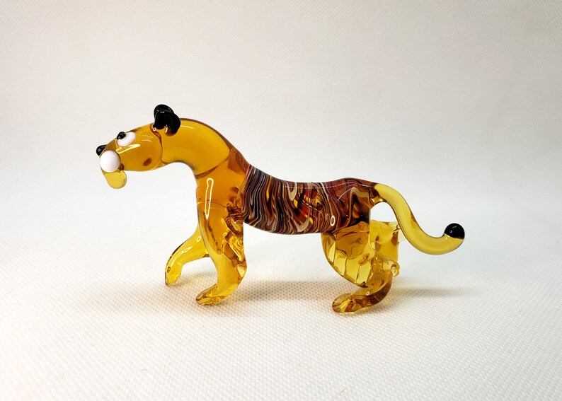 K&ouml;nnte beinhalten: Eine Glasfigur eines Tigers mit einem gelb-braun gestreiften K&ouml;rper, schwarzen Ohren und wei&szlig;en Augen. Die Figur steht auf einer wei&szlig;en Oberfl&auml;che.