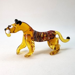 K&ouml;nnte beinhalten: Eine Glasfigur eines Tigers mit bernsteinfarbenen und braunen Streifen. Der Tiger hat schwarze Augen und eine wei&szlig;e Nase.