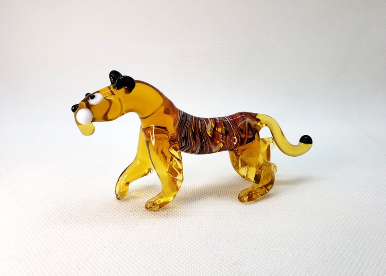K&ouml;nnte beinhalten: Eine Glasfigur eines Tigers mit bernsteinfarbenen und braunen Streifen. Der Tiger hat schwarze Augen und eine wei&szlig;e Nase.