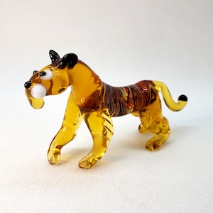 K&ouml;nnte beinhalten: Eine Glas-Tigerfigur mit bernsteinfarbenen und braunen Streifen. Der Tiger hat schwarze Augen und einen wei&szlig;en Fang.