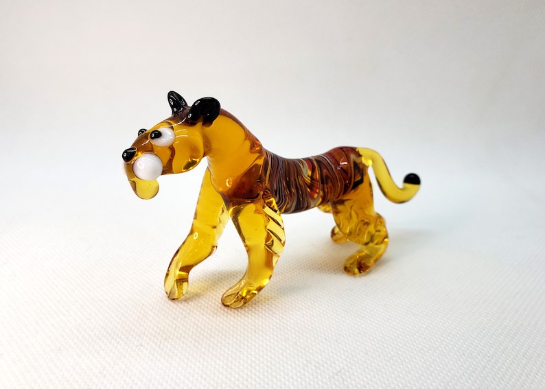 K&ouml;nnte beinhalten: Eine Glas-Tigerfigur mit bernsteinfarbenen und braunen Streifen. Der Tiger hat schwarze Augen und einen wei&szlig;en Fang.