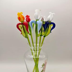May include: A collection of handcrafted glass flowers in a clear glass vase. The flowers feature vibrant petals in red, yellow, white, and blue, each attached to a long green glass stem with green leaves. The vase has a floral decal on the side.