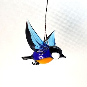 Puede incluir: Un adorno de pájaro de vidrio azul y naranja con alas negras y un ojo blanco. El pájaro está en vuelo y tiene un colgador de alambre.