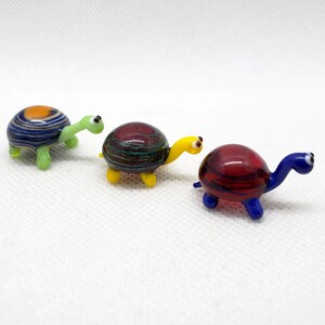 May include: Three small glass turtle figurines. The turtles are each a different colour: blue, green, and yellow. The turtles have striped shells and black eyes.