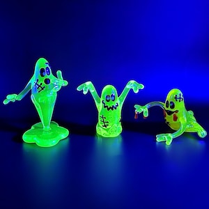 May include: Three glowing green ghost figurines with black stitched details and cartoon faces. One stands on a base, another has arms raised, and the third has a red drop. Set against a dark blue background.
