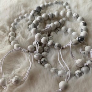 May include: Several white and gray beaded bracelets with macrame knots and white string. The beads have a marbled appearance. The bracelets are arranged on a fluffy, white surface.