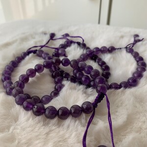 May include: Several amethyst bead bracelets are displayed on a white, fluffy surface. Each bracelet is made of round, polished purple beads strung on a purple cord. The bracelets are adjustable, with a sliding knot closure. The beads have a translucent quality.