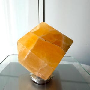 May include: A faceted, golden-yellow calcite crystal, resting on a small, circular, silver-colored base. The crystal's surface has natural variations and internal veining. The object is displayed on a reflective, gray surface.