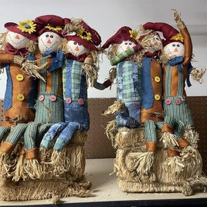 May include: Two decorative sets of three fabric scarecrows sitting on rectangular straw bales. Each scarecrow features a round white face, a red hat with a yellow sunflower, and colorful patterned clothing with buttons, straw hair, and hands.