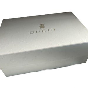 以下が含まれることがあります： ブランド名「GUCCI」が金色で書かれた白い長方形の箱。箱には繰り返しの模様と小さなグラフィックがあります。箱は閉じており、段ボールでできているようです。