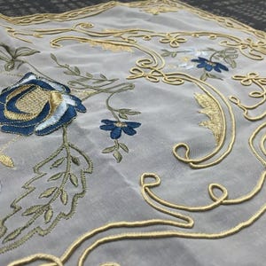 May include: A white sheer tablecloth with gold and blue embroidered floral designs. The tablecloth features a large blue rose with gold accents, smaller blue flowers, and gold swirling patterns.