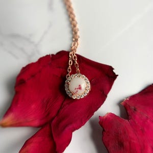 May include: A delicate rose gold necklace with a white pendant featuring a pink and white floral design. The pendant is surrounded by small clear stones.