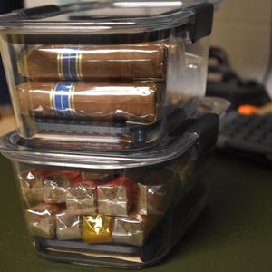 May include: Two clear plastic containers stacked on top of each other. The top container holds two cigars, wrapped in brown paper with blue and gold bands. The bottom container holds several small, individually wrapped candies.