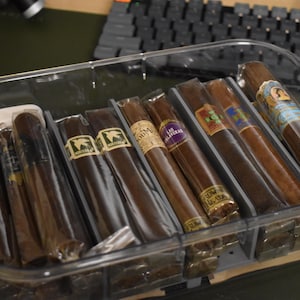 May include: A clear plastic container filled with several cigars. The cigars are various shades of brown and are individually wrapped. Some cigars have colorful bands with text and designs. The container sits on a green surface.