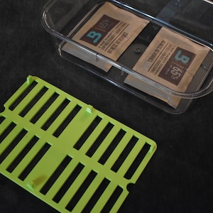 May include: A clear plastic container holding two brown packets with white text. A bright green slotted tray is in the foreground. The items are on a dark surface.
