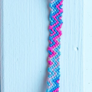 May include: A friendship bracelet made with pink, light blue, and dark blue embroidery floss. The bracelet is tied in a knot at the top and has a chevron pattern.