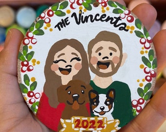 May include: A round wooden ornament with a white background and a red, green, and gold wreath border. The ornament features a hand-painted portrait of a couple with two dogs, the text "The Vincenta" and the year "2022".
