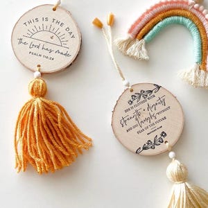 May include: Wooden disc ornaments with inspirational quotes and tassels. One ornament reads "This is the day the Lord has made" and the other says "She is clothed with strength and dignity". A rainbow decoration is also visible.