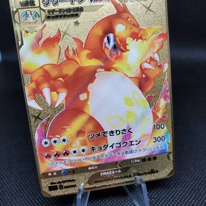 May include: A gold Pokemon trading card featuring a holographic image of a red and orange Charizard VMAX with the text "VMAX リザードン VMAX HP 330" and "ツメできりさく 100 キョダイゴクエン 300" on the card.