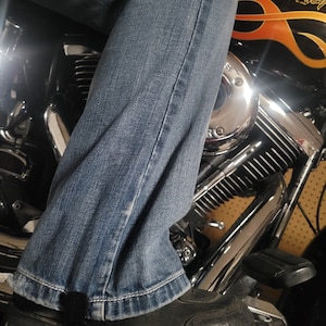 May include: A person wearing blue jeans and black boots sits on a motorcycle with a black and orange paint job. The motorcycle has a chrome engine and exhaust pipes. The motorcycle has the word "Harley" written on the side.