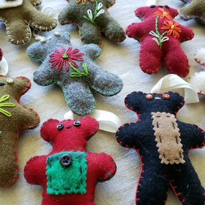May include: A collection of felt teddy bear ornaments with various embroidered details, including flowers, buttons, and patches. The ornaments are in shades of brown, green, red, black, and white.