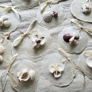 May include: A collection of 10 white seashell ornaments with various seashells and starfish glued to the front. The ornaments are tied with white ribbon and twine.