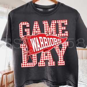 May include: A charcoal grey short-sleeve t-shirt featuring the text GAME DAY in a red and white gingham pattern. A red pennant graphic is layered over the text with the word WARRIORS in white, accented by two red bows on the left side.