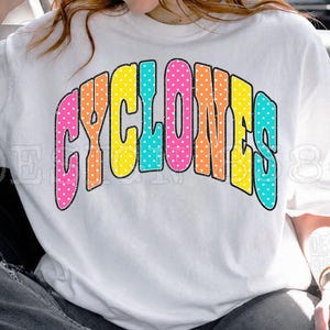 Può includere: Una t-shirt bianca a maniche corte con la parola CYCLONES stampata in un grande carattere arcuato e multicolore. Ogni lettera ha un colore distinto, tra cui rosa, arancione, giallo e verde acqua, decorata con un motivo a piccoli puntini bianchi.