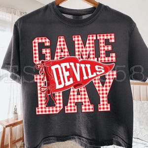 May include: A charcoal gray short-sleeve t-shirt featuring the text GAME DAY in a red and white gingham pattern. A red pennant graphic with white text reading DEVILS is layered over the center, accented by two red bows on the left side.