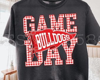May include: A charcoal grey short-sleeve t-shirt featuring the text GAME DAY in a red and white gingham pattern. A red pennant graphic with the word BULLDOGS in white letters and two red bows is layered over the centre of the text design.