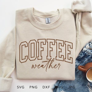 May include: A beige sweatshirt with the words "COFFEE weather" in brown lettering. The sweatshirt is folded and placed on top of a pair of blue denim shorts and a brown mug filled with coffee.