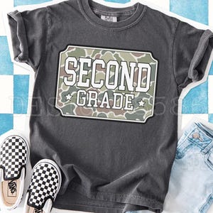 May include: A charcoal gray short-sleeve t-shirt featuring a rectangular graphic with a camouflage pattern. The text SECOND GRADE is printed in white block letters inside the rectangle, flanked by two small white stars on the bottom row.