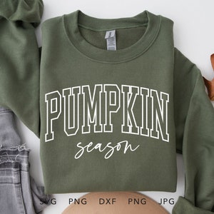 May include: An olive green sweatshirt featuring the text "PUMPKIN season" in white. The sweatshirt is folded, with grey jeans and a fallen leaf in the background. The sweatshirt is made of a soft material, perfect for autumn.